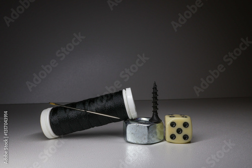 A skein of black threads with a needle rests on a nut, a black self-tapping screw and a dice