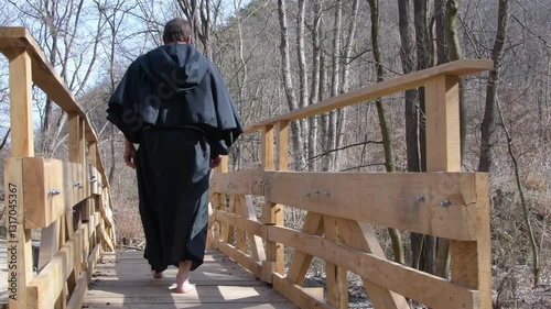 A figure in a dark cloak and hood walks across a bridge in a leafless forest. 