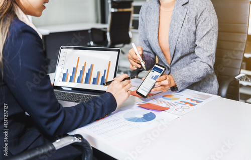Two female business professionals in suits sit at desk, engaged in strategic discussion. They analyze financial planning, risk management, corporate growth while reviewing reports and market trends.