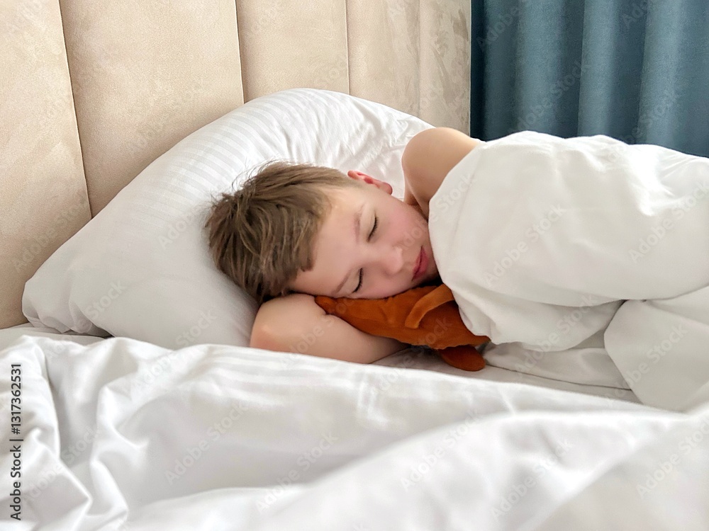 A young boy sleeps peacefully on a plush bed, nestled under a soft white blanket, hugging a small stuffed toy A young boy sleeps peacefully on a plush bed, nestled under a soft white blanket, hugging a small stuffed toy