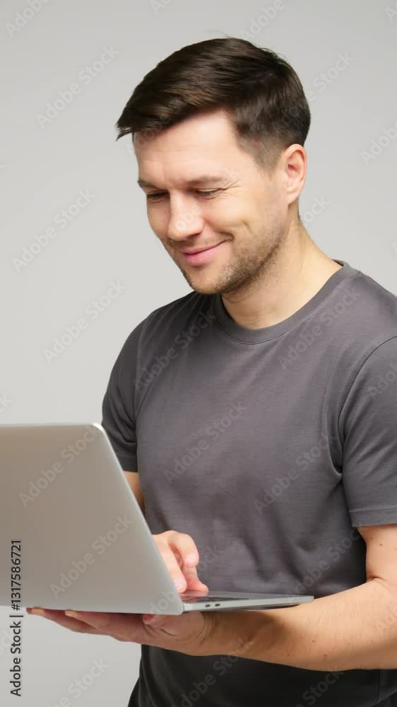 Smiling young man holding laptop while standing against neutral background in modern indoor space Smiling young man holding laptop while standing against neutral background in modern indoor space