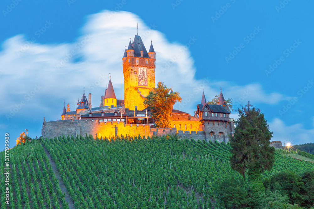 Reichsburg Castle on a hill surrounded by vineyards with illuminated buildings and a cloudy sky at dusk in Cochem, Germany