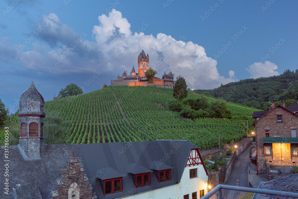 Reichsburg Castle on a hill surrounded by vineyards with illuminated buildings and a cloudy sky at dusk in Cochem, Germany