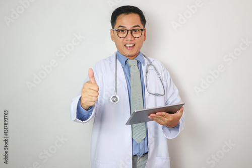 A handsome, smiling Asian male doctor in a white gown uniform showing thumb up while using digital tablet, reading medical cases, standing against on a white studio background.