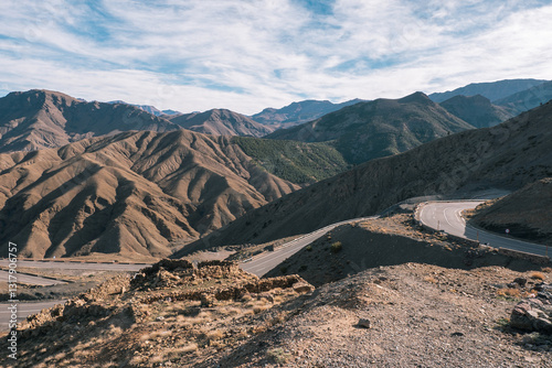 A winding road cuts through the rugged landscape of the Atlas Mountains in Morocco, under a partly cloudy sky.