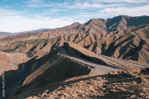 A winding road cuts through the rugged landscape of the Atlas Mountains in Morocco, under a partly cloudy sky.