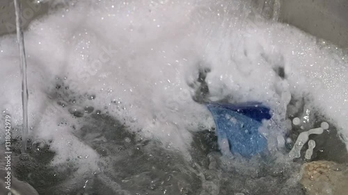 Kitchen sink filling with soapy water to hand wash dishes.  Closeup view of household chores in slow motion.  Soap suds build as faucet splashes and fills sink.
