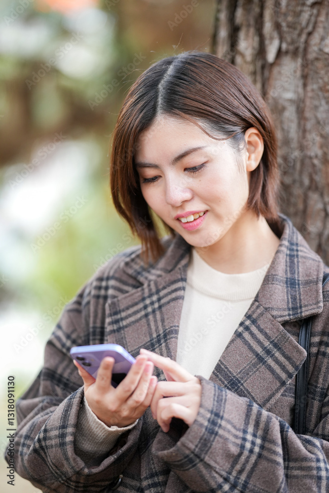 A 29-year-old Japanese woman wearing a checkered coat. She smiles and operates her smartphone next to the tree. A 29-year-old Japanese woman wearing a checkered coat. She smiles and operates her smartphone next to the tree.