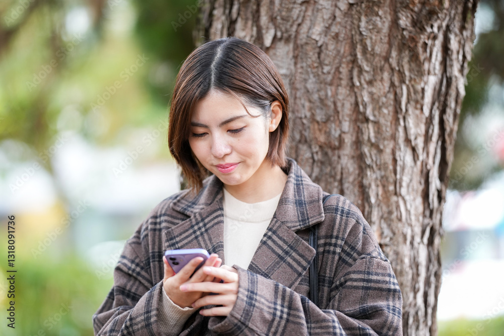 A 29-year-old Japanese woman wearing a checkered coat. She smiles and operates her smartphone next to the tree. A 29-year-old Japanese woman wearing a checkered coat. She smiles and operates her smartphone next to the tree.