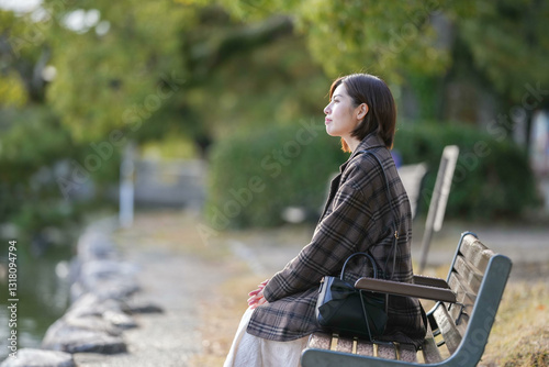 Winter at Ohori Park in Chuo Ward, Fukuoka City, Japan. A 29-year-old Japanese woman wearing a checkered coat. She is sitting on a bench along the pond. photo