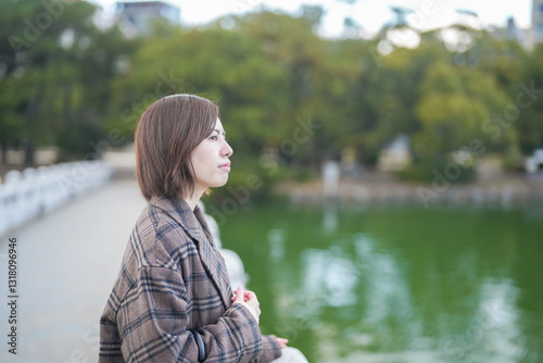 Winter at Ohori Park in Chuo Ward, Fukuoka City, Japan. A 29-year-old Japanese woman wearing a checkered coat. She warms her hands together to get out of the cold, looking out at the pond from the bri