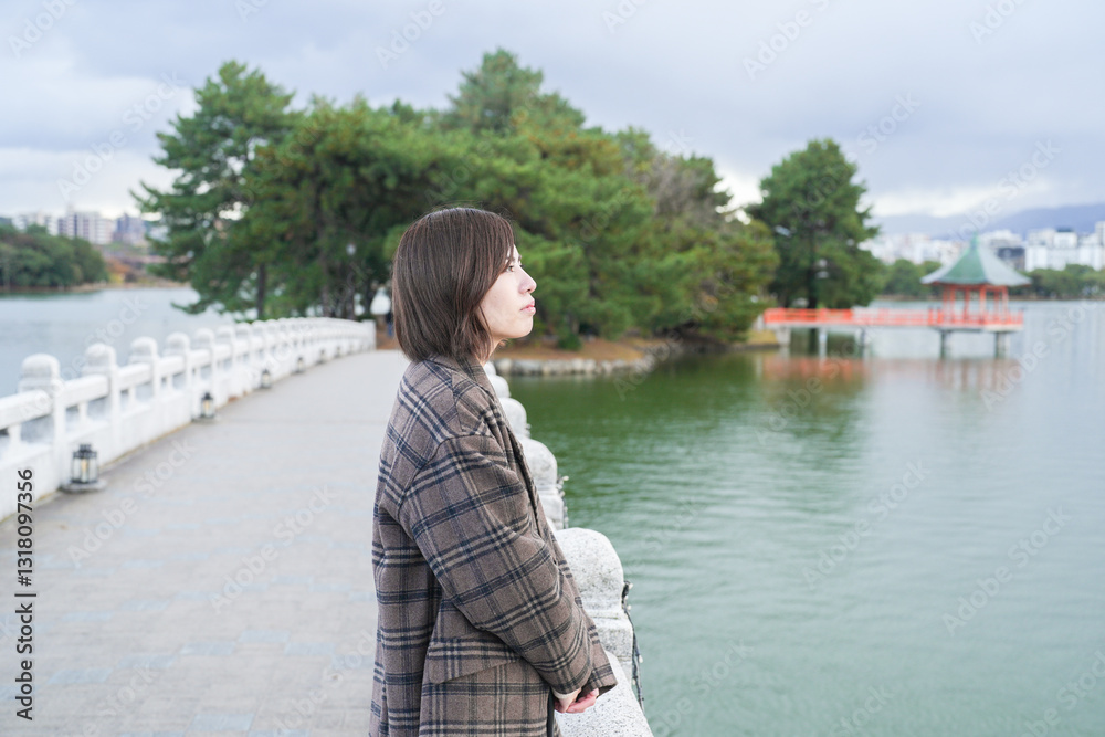 Winter at Ohori Park in Chuo Ward, Fukuoka City, Japan. A 29-year-old Japanese woman wearing a checkered coat. She warms her hands together to get out of the cold, looking out at the pond from the bri Winter at Ohori Park in Chuo Ward, Fukuoka City, Japan. A 29-year-old Japanese woman wearing a checkered coat. She warms her hands together to get out of the cold, looking out at the pond from the bri