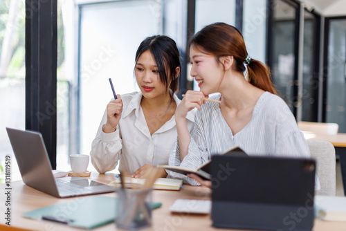 Happy professional young women collaborating with enthusiasm in a modern office setting. Two asian female entrepreneurs using a laptop for a productive business meeting in a corporate workplace.
