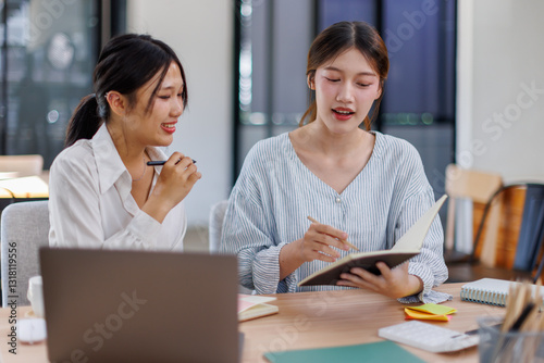 Happy professional young women collaborating with enthusiasm in a modern office setting. Two asian female entrepreneurs using a laptop for a productive business meeting in a corporate workplace.
