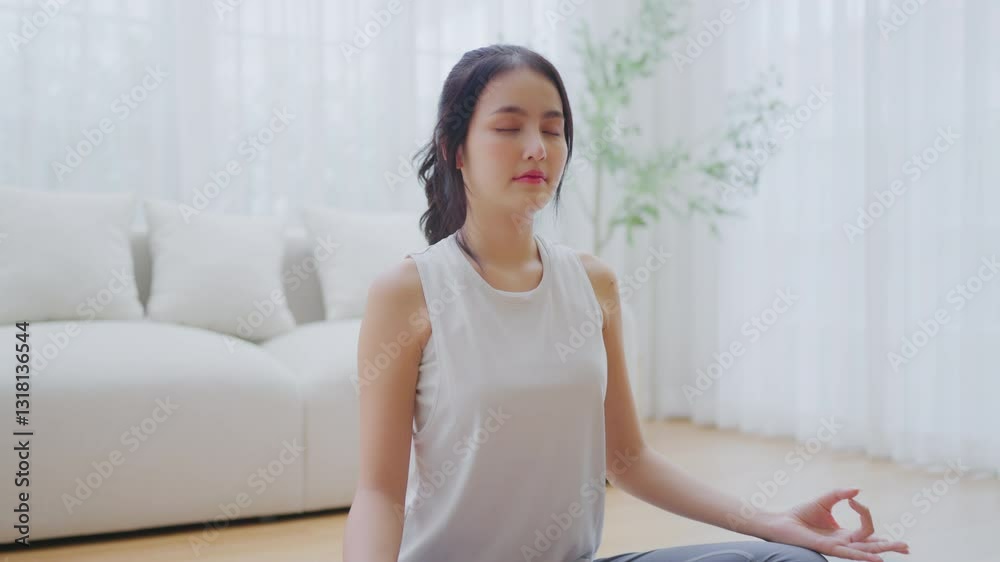 Happy young Asian woman practicing yoga and meditation at home sitting on floor in living room in lotus position and relaxing with closed eyes. Mindful meditation and wellbeing concept Happy young Asian woman practicing yoga and meditation at home sitting on floor in living room in lotus position and relaxing with closed eyes. Mindful meditation and wellbeing concept