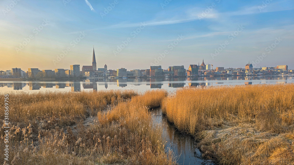 Fototapeta premium scenic view of the city skyline of Rostock, germany, reflecting on a calm river at sunset. A small waterway winds through golden reeds in the foreground
