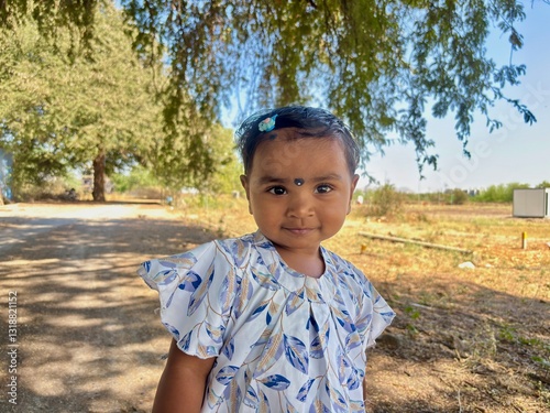 Portrait of a 2-year-old Asian girl standing in the shade under a tree, smiling slightly with a hint of shyness while looking at the camera.