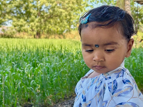 A 2-year-old Asian girl stands outdoors with her body facing forward while turning her head over her shoulder, looking down at something on the ground with a curious expression.
