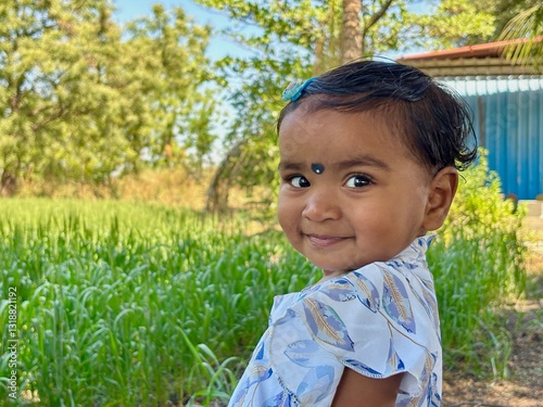 A 2-year-old Asian girl stands with her body facing forward while turning her head over her shoulder to look at the camera, smiling brightly.