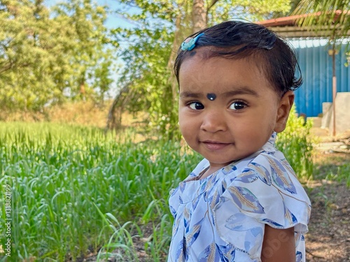 A 2-year-old Asian girl stands with her body facing forward while turning her head over her shoulder, looking at the camera with a slight smile.