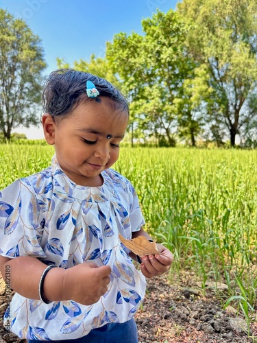 Portrait of a 2-year-old Asian girl standing on a farm, holding an old leaf from a tree and gently smiling while looking at it.
