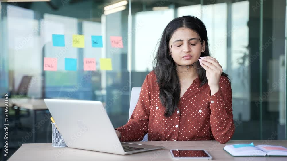 Tired young businesswoman suffering from eye strain while working on a laptop sitting at desk at a workplace in a business office. Overworked woman takes off glasses and massages, rubs bridge of nose