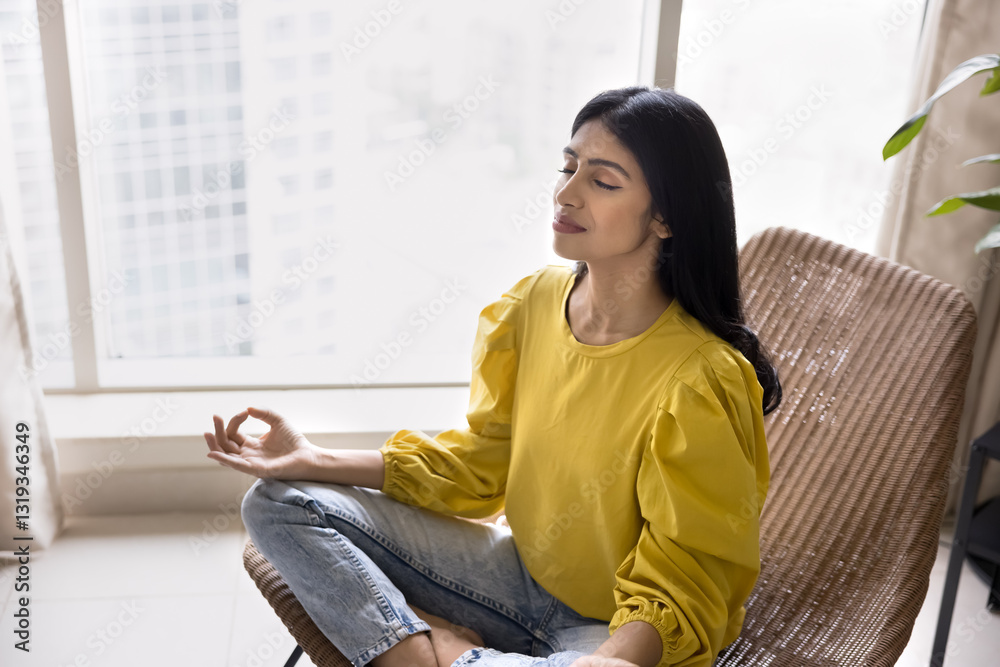 Fototapeta premium Young Indian woman doing meditation practice in modern high-rise building apartment, relaxing in hustle and bustle of city life, reduces symptoms of anxiety and worry, strengthen mind-body connection