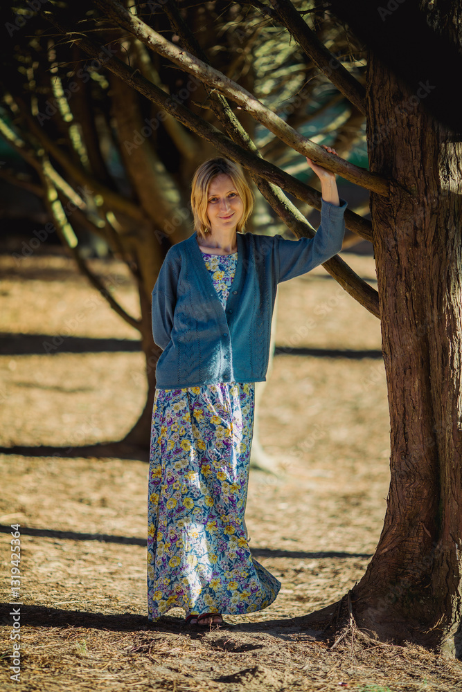 A woman in a long summer dress stands near a tree in a park during spring.