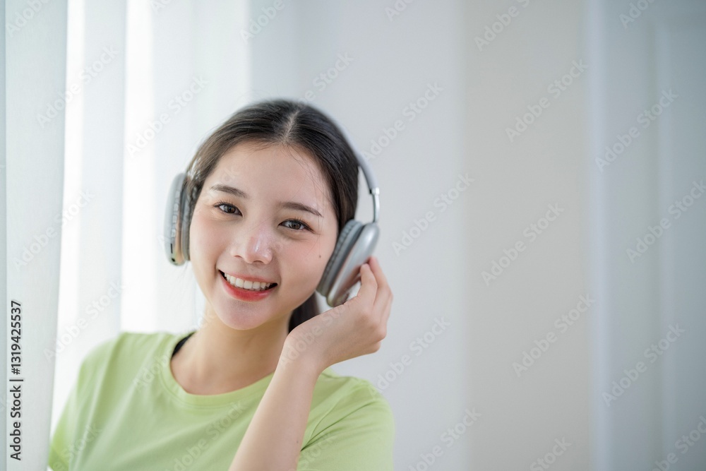 Naklejka premium Young woman enjoys music while wearing headphones and smiling in a bright, airy room during daytime