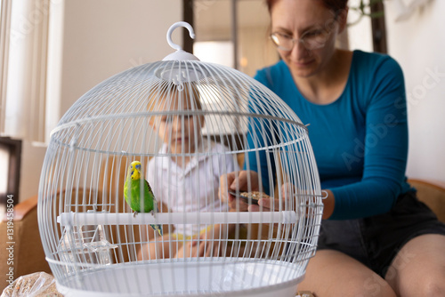 Mother and child feeding a green budgie in a cage