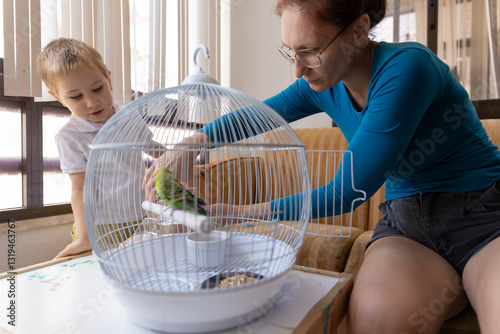 Mother and son caring for pet budgie in cage at home