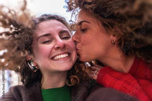 Two cheerful young women are enjoying time together, with one giving a kiss on the cheek to the other