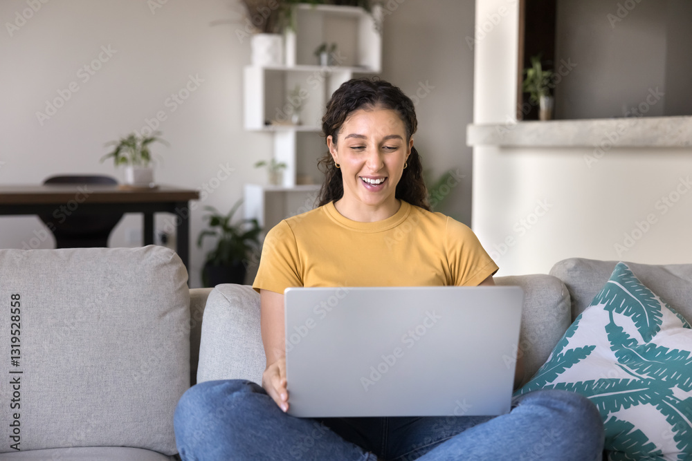 Naklejka premium Young Latina woman laughing while watching online movie via subscription streaming services, resting on couch with modern laptop, enjoy videos and on-line content spend carefree pastime alone at home