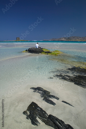 Man sitting on a rock, beach and rocks. La Pelosa, Stintino, Sassari, Sardinia, Italy