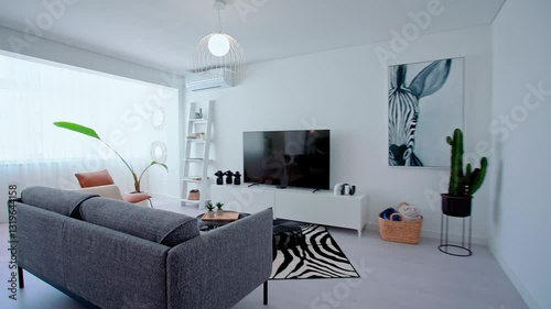A contemporary living room features a gray sofa with decorative pillows, a zebra-striped rug, a round coffee table, and a brown leather chair near a large window with sheer curtains.