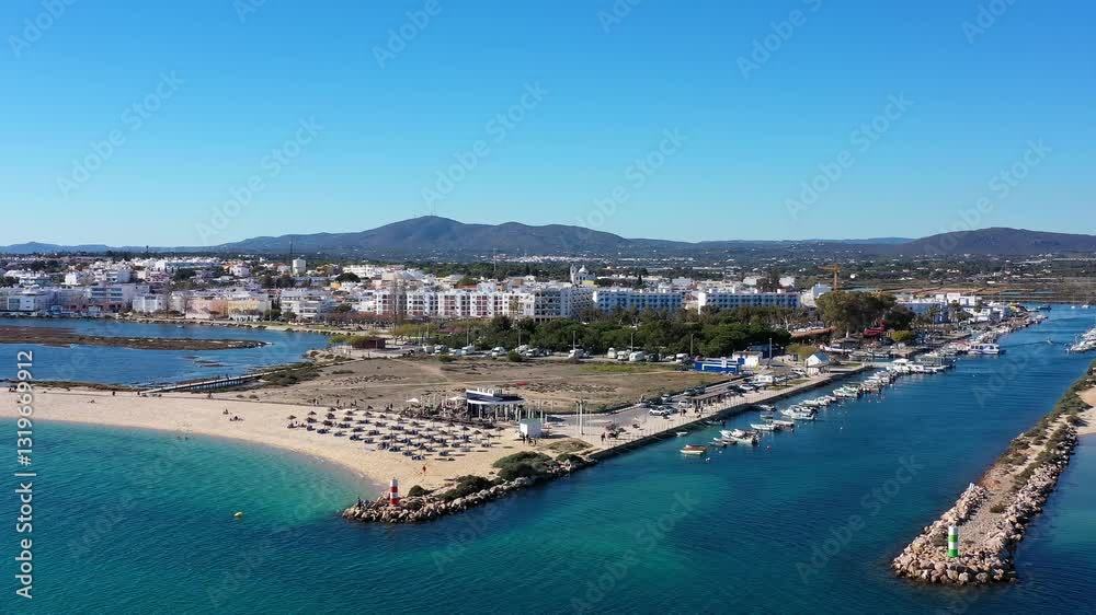 Aerial view of Fuseta Portugal showcasing a sandy beach lined with sun umbrellas a marina filled with boats a picturesque channel leading to Ria Formosa and distant mountains Aerial view of Fuseta Portugal showcasing a sandy beach lined with sun umbrellas a marina filled with boats a picturesque channel leading to Ria Formosa and distant mountains