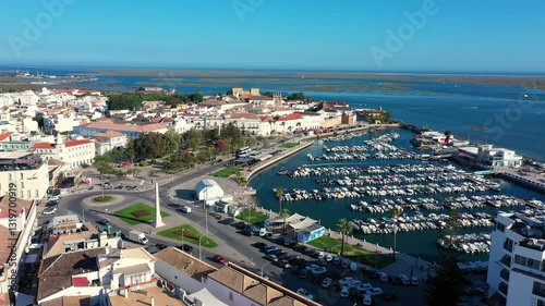 Aerial view of Faro, Portugal, showcasing the historic city center, marina filled with boats, green park areas, and the stunning blue waters of the Ria Formosa lagoon stretching to the horizon.