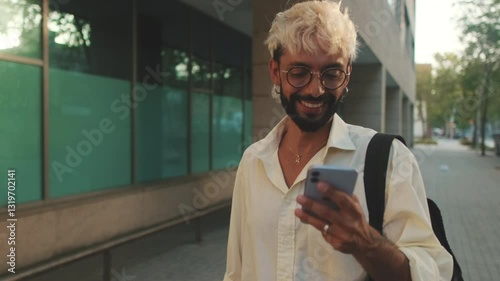 A young man with bleached blond hair and a beard smiles while walking down a city sidewalk and looking at his phone.