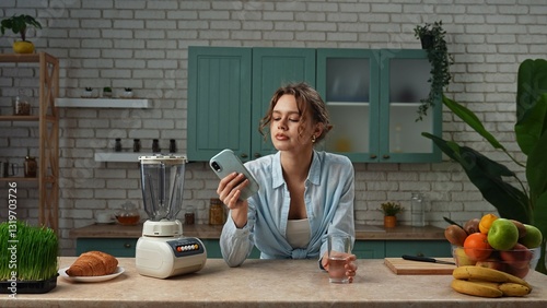 Young woman standing at the table in the kitchen and preparing healthy breakfast, drinking clear water from glass, scrolling media on smartphone.