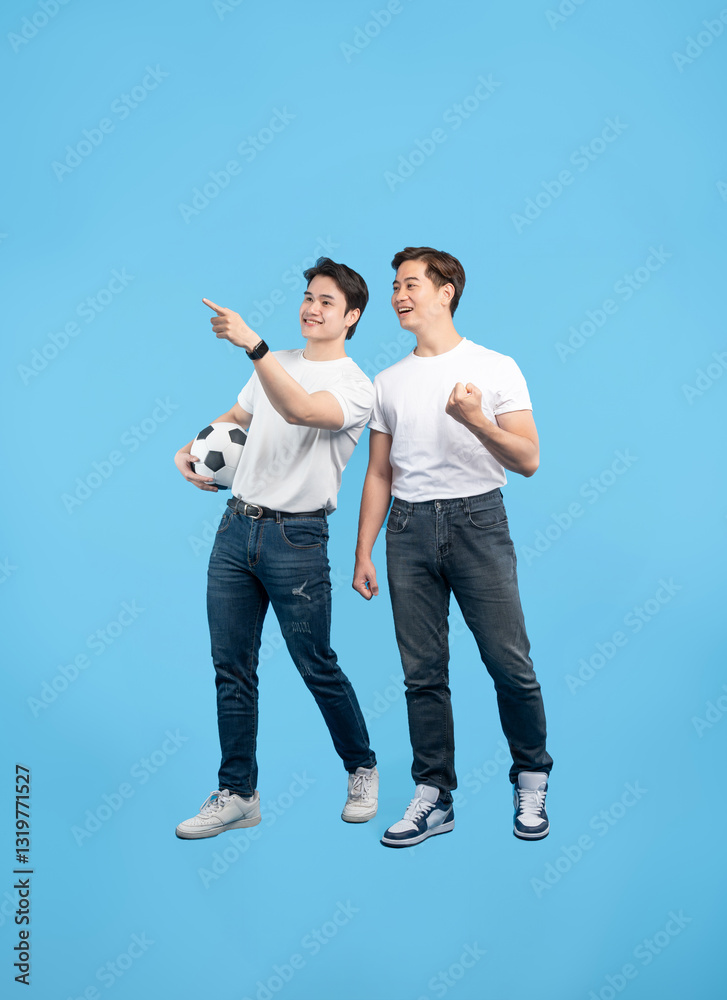 Fototapeta premium Two friends anticipating a soccer match, one holding a ball and pointing, both in casual attire against a blue background. Excitement visible.