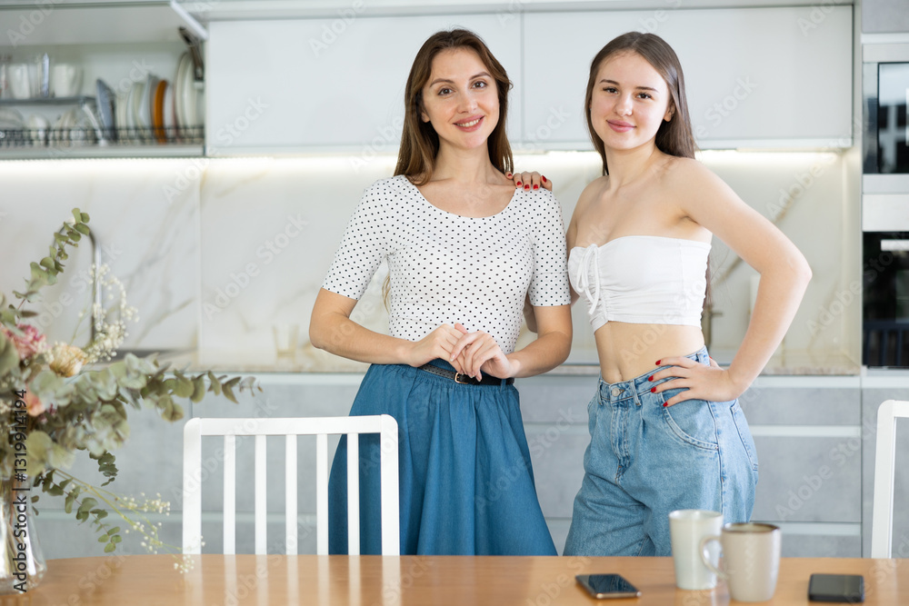 Joyous mother and her teenage daughter standing together against the background of white kitchen cabinet Joyous mother and her teenage daughter standing together against the background of white kitchen cabinet