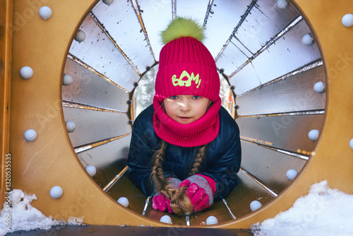 A young girl crawls through a playground tunnel covered in snow, wearing a bright winter hat and gloves, enjoying her time outdoors