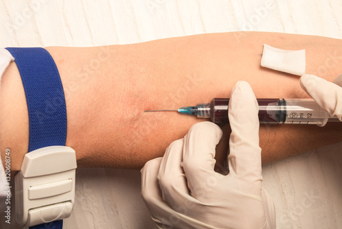 A healthcare professional wearing latex gloves draws blood from a patient's arm using a syringe and needle. A tourniquet is applied and a cotton swab is prepared on a white background