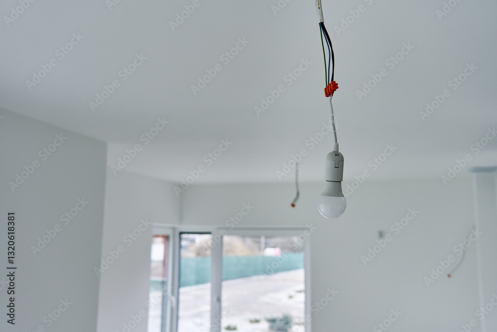 Single light bulb hanging from exposed electrical wires in unfinished interior with white walls and concrete flooring. Lighting in building under renovation
