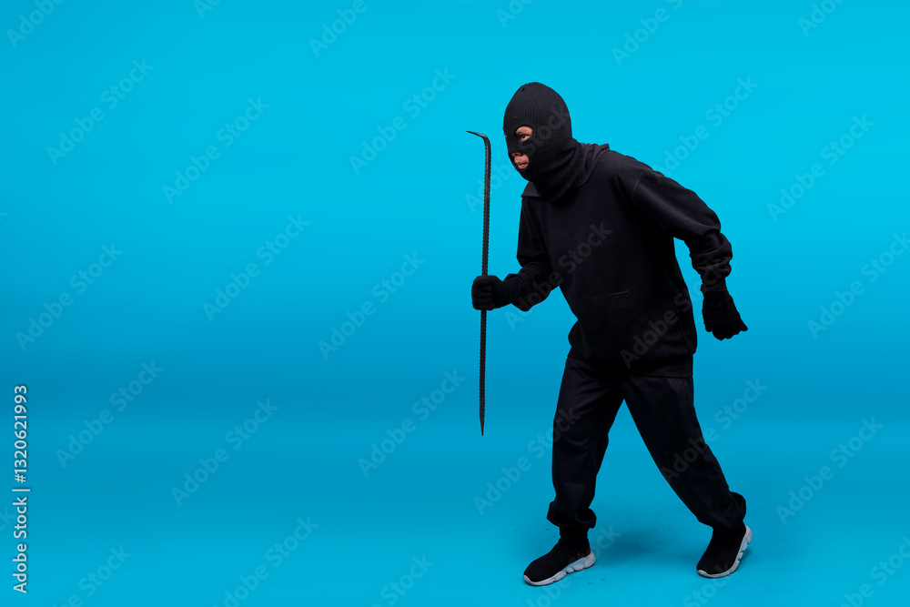 Fototapeta premium Robber In Black Clothes Wearing Walking While Holding Crowbar Isolated On Blue Background