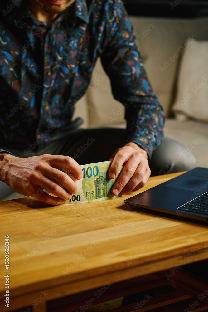 Man holding one hundred euro banknotes, sitting on sofa in front of table with open laptop. Person counting cash money at home. Personal finance and home budget concept