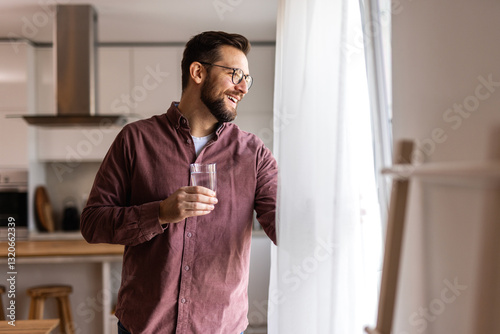A man in casual wear drinks water in a modern kitchen, taking a moment to recharge. He stays hydrated to maintain focus, wellness, and productivity in his daily lifestyle.