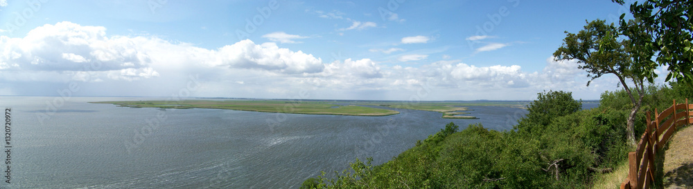 Beautiful wide landscape - blue lake in evergreen forest under fluffy clouds in the sky on a sunny day, with green grass in the foreground