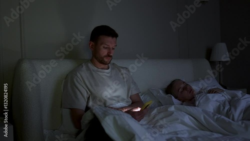 Cheating husband scrolls phone, chats on social networks, messaging in bed at night. Wife sleeps while man betrays absorbed in smartphone, web feed and digital addiction, irregular work schedule