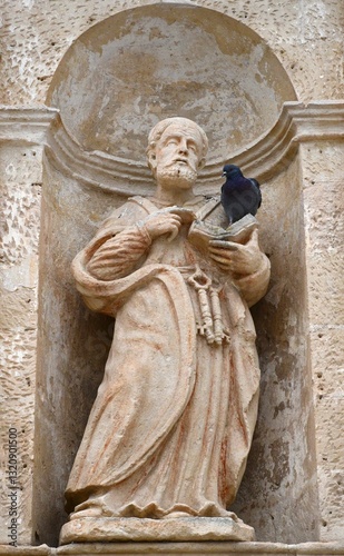 An old stone statue of St Peter stands in the niche of a wall. His symbolic keys hang from his belt. He holds an open book, where a pigeon is resting. The stone is pale, with faded red paint on St Pet